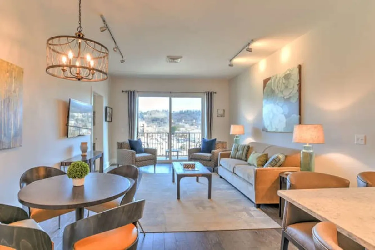 The image showcases a spacious and elegantly furnished living and dining area filled with natural light from a large window revealing sunrise views over downtown Asheville. A plush sofa invites relaxation, while a modern chandelier hangs above a dining table set for intimate meals, creating a warm and inviting ambiance perfect for socializing and celebrating with family or friends.