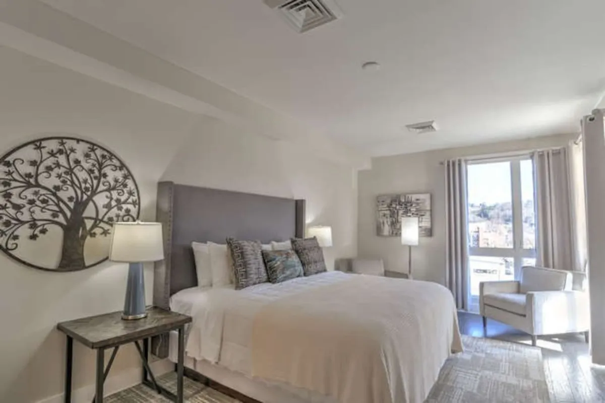 A spacious bedroom featuring a large bed with plush bedding, a decorative tree wall art above the headboard, stylish bedside lamps, a comfortable armchair, and a window streaming in natural light. The neutral palette and soft textures create an inviting, serene atmosphere ideal for relaxation and quiet moments.
