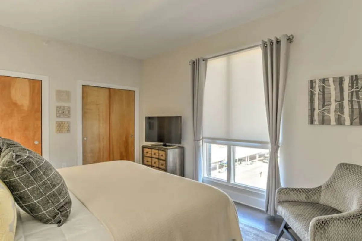 This image showcases a stylish bedroom featuring a cozy bed adorned with decorative pillows, a television unit, a window with soft curtains, and tasteful wall art. The room is filled with inviting morning light, creating a welcoming and restful ambiance ideal for unwinding after a busy workday.
