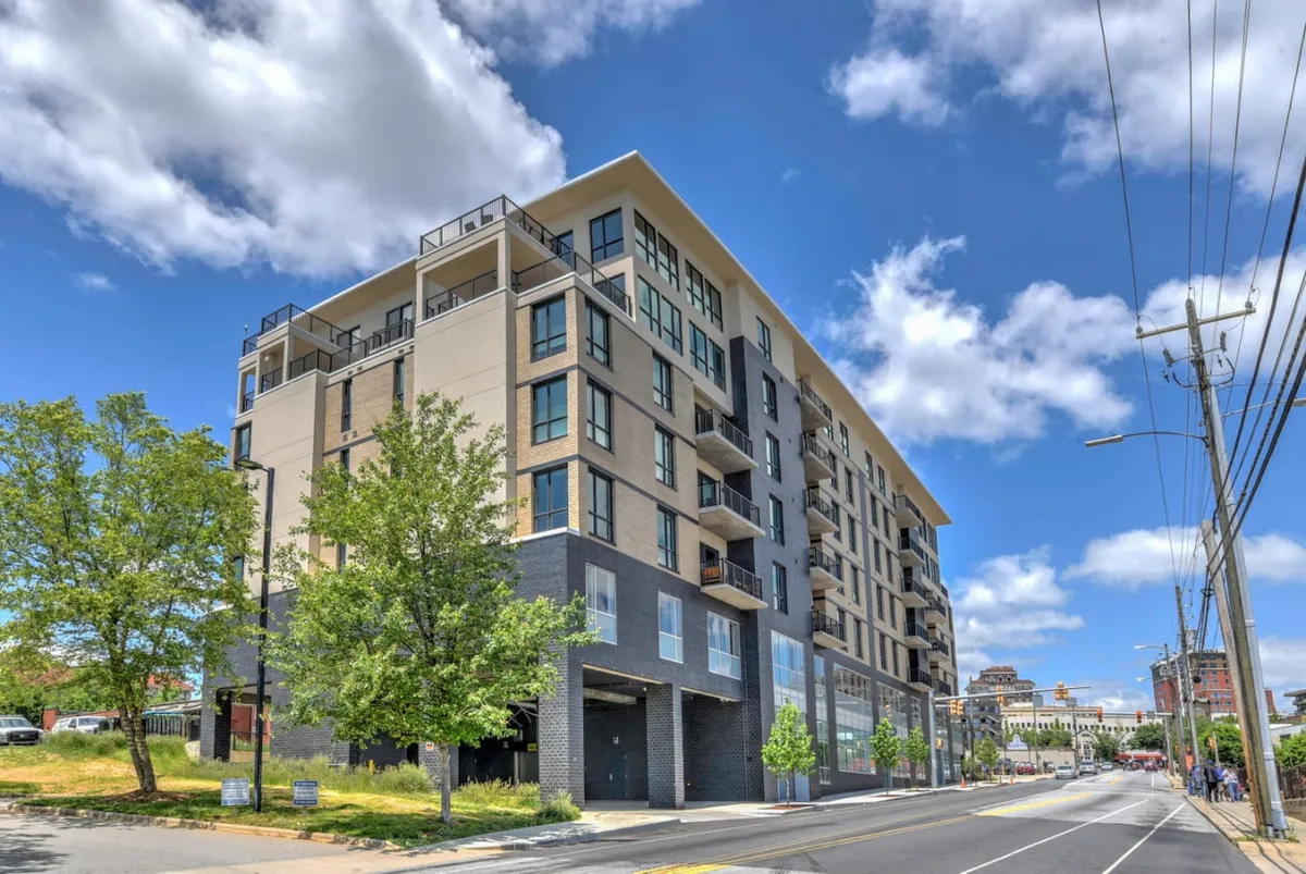 This image highlights the modern exterior of the condo building, surrounded by green trees and set against a clear sky. The scene is lively and contemporary, with hints of rooftop city views and a sense of community gathering, showcasing the building's proximity to Asheville's vibrant downtown attractions.