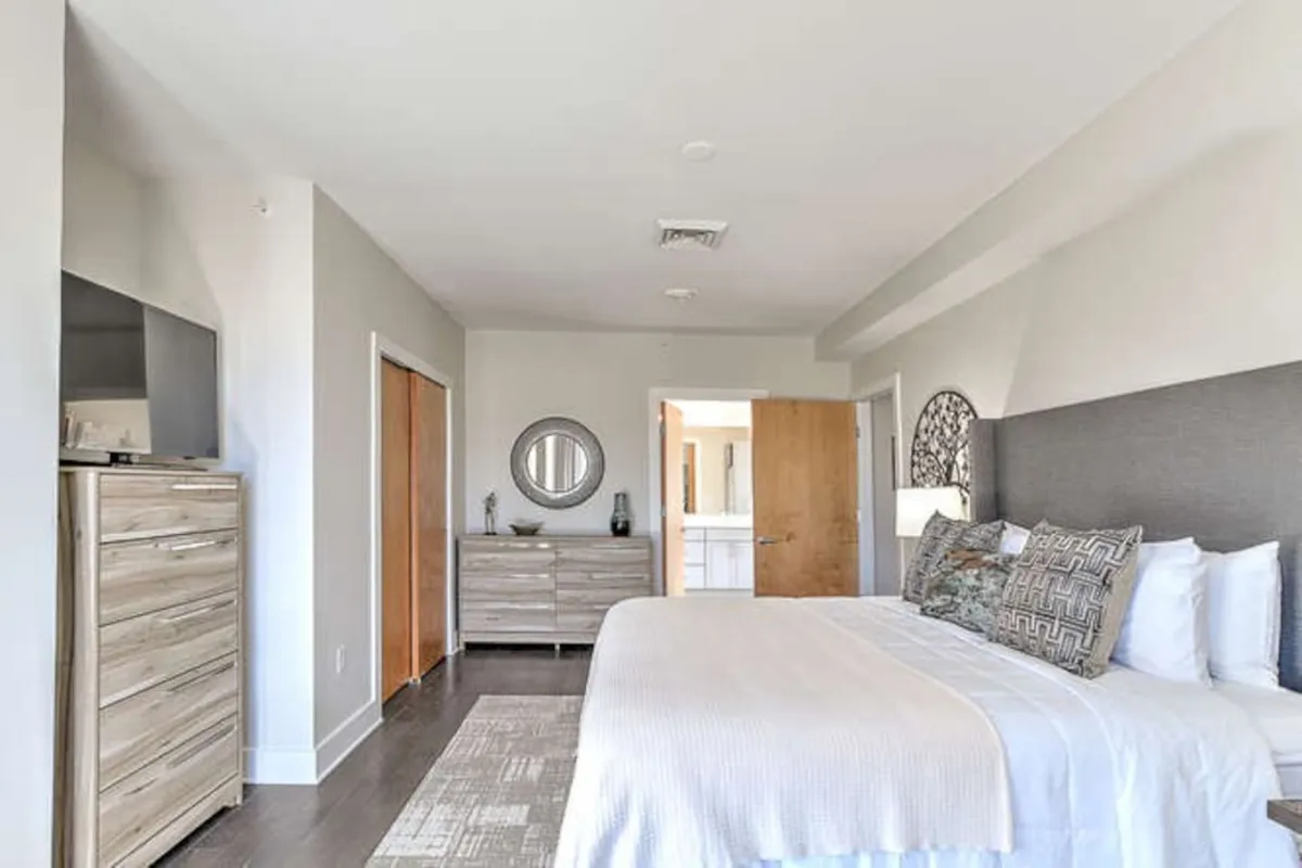A welcoming bedroom scene highlighting a bed adorned with decorative pillows, a dresser, a round mirror, and a visible entrance to a private bathroom. Sunlight filters in, amplifying the room's serene and comfortable vibe. The bathroom door in view connects the concept of private en-suite bathrooms.