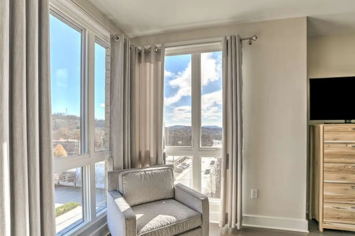 A cozy armchair is positioned near large windows, with soft curtains framing an urban view. The scene includes a dresser and gentle daylight filtering in, creating a relaxing space that subtly hints at the condo's connection to the lively city outside.