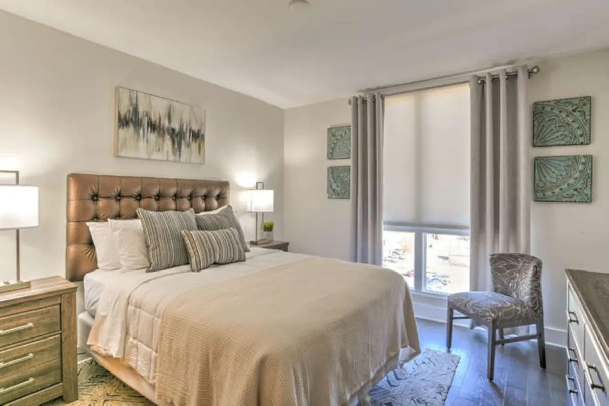 A tranquil bedroom scene featuring a plush bed with a tufted headboard, decorative pillows, a softly glowing nightstand lamp, and tasteful artwork. The lighting and arrangement evoke a sense of peaceful privacy ideal for couples seeking a romantic retreat.