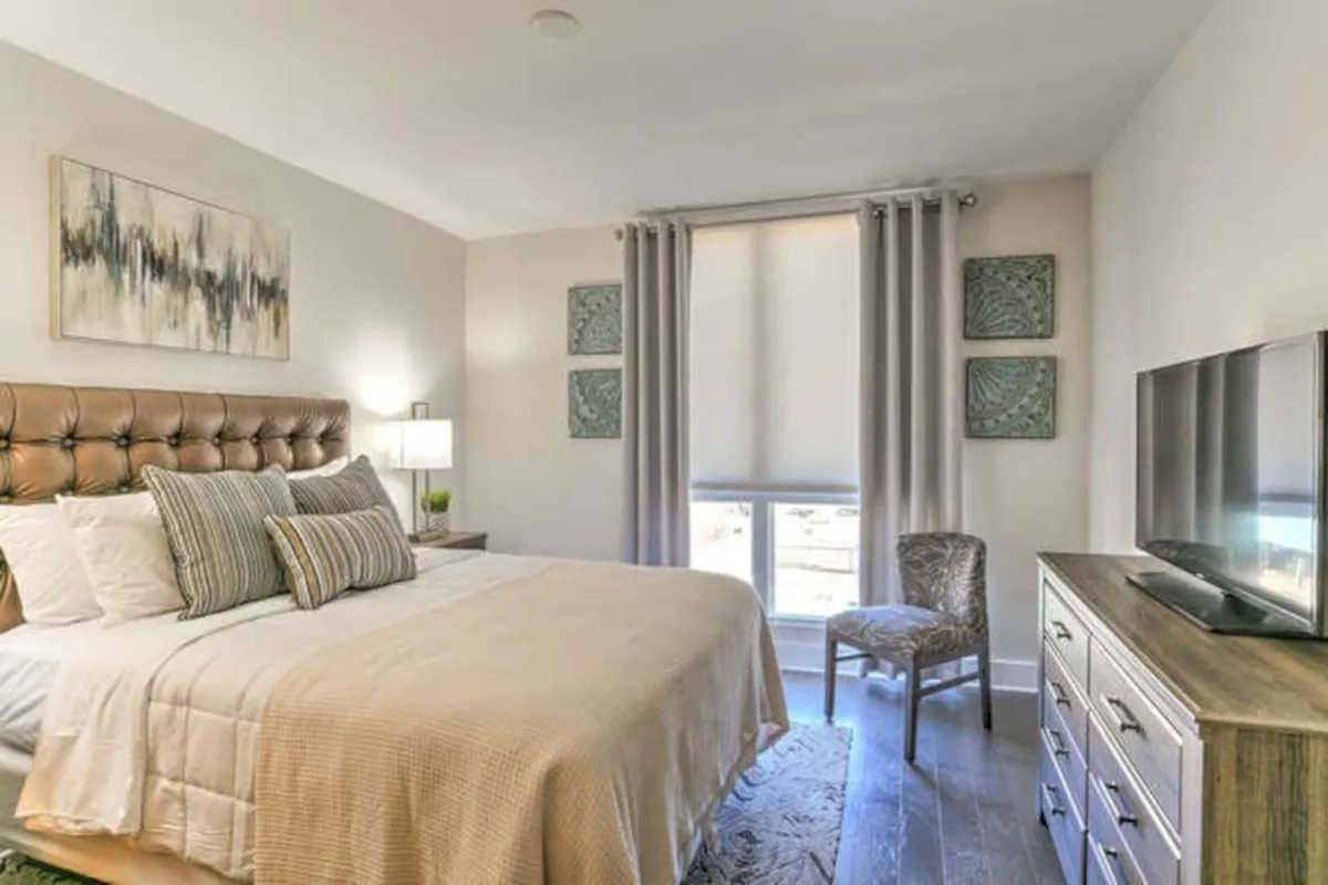 A cozy bedroom features a tufted headboard, elegant wall art, a TV atop a dresser, and a comfortable chair by the window. Natural light streams in, highlighting the room’s inviting atmosphere for quiet moments or cozy evenings in.