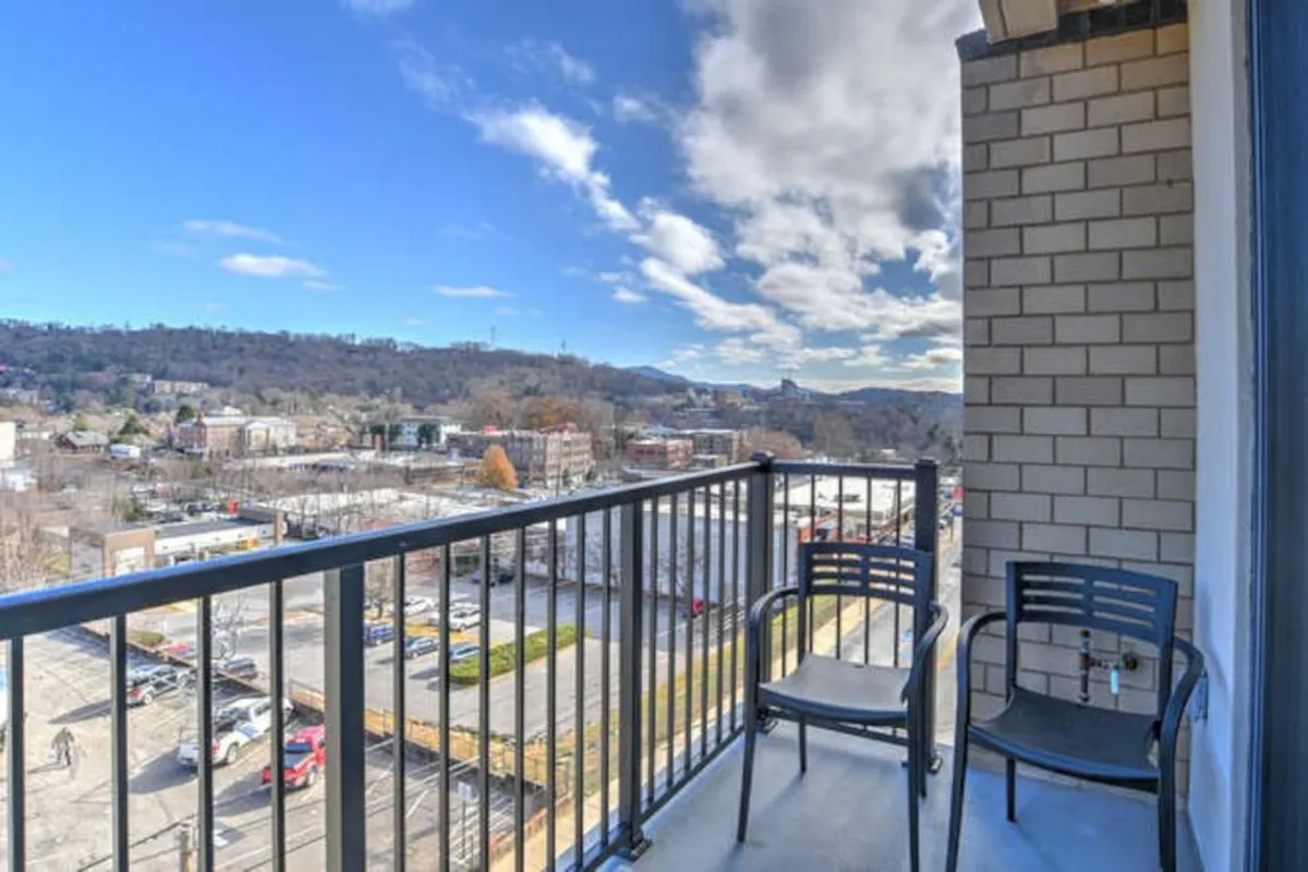 A serene private balcony furnished with two chairs and a sleek railing, offering a breathtaking panoramic view of the city skyline. The scene hints at tranquil moments, whether enjoying a morning coffee or unwinding at sunset, just steps from the vibrant living area inside.