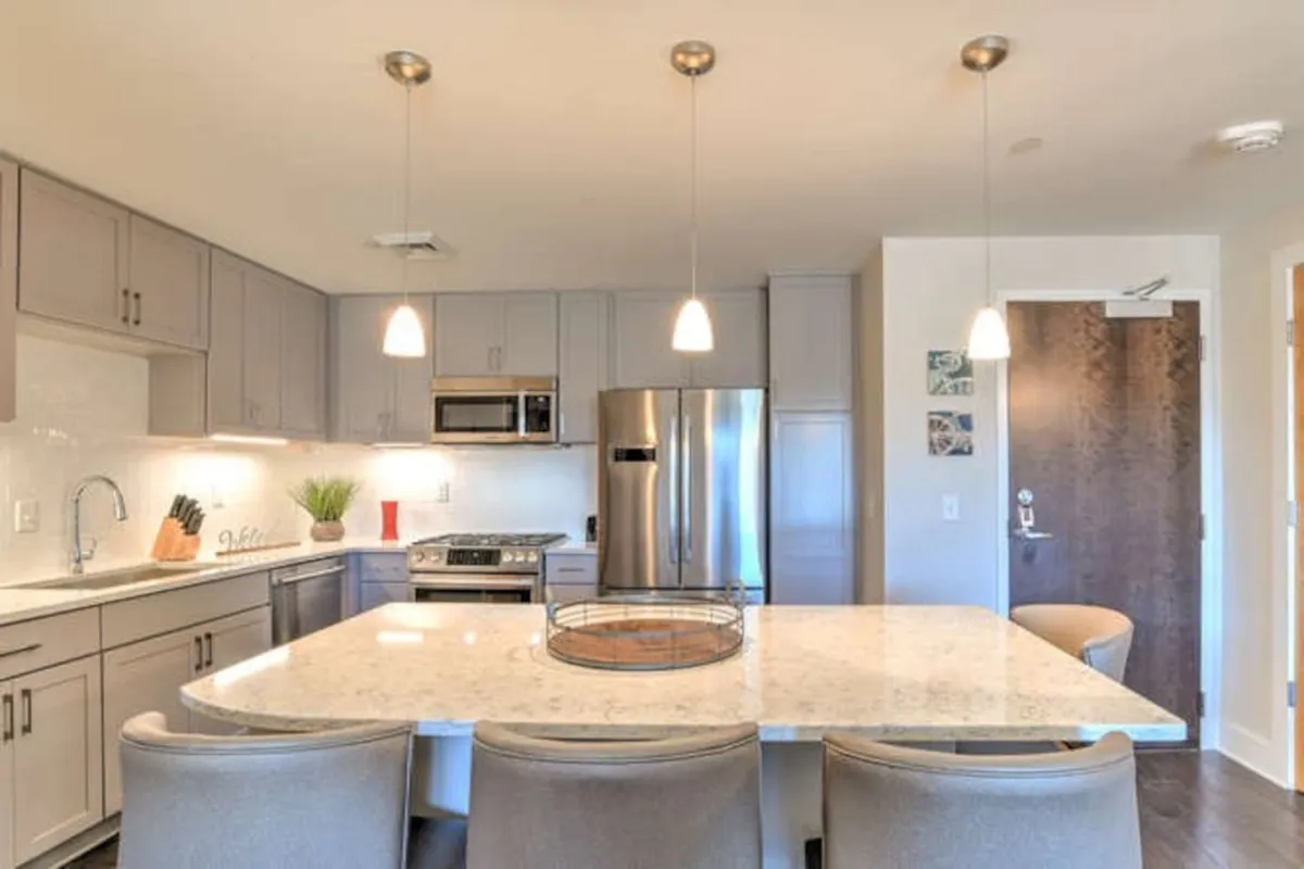 This image shows a modern kitchen with a spacious island countertop, stainless steel appliances, and stylish pendant lighting. The arrangement makes the kitchen inviting for both cooking and casual gatherings, with ample prep space and a door leading to another area, suggesting an open and connected layout.