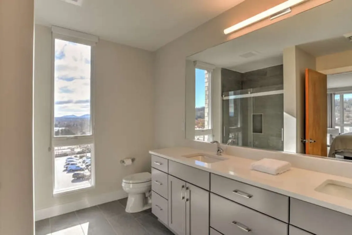 A modern, serene bathroom featuring dual sinks, a large mirror, a modern shower, and a window that lets in natural light. The clean lines and spacious countertop create an atmosphere of relaxation and freshness—ideal for personal routines and unwinding after a busy day.